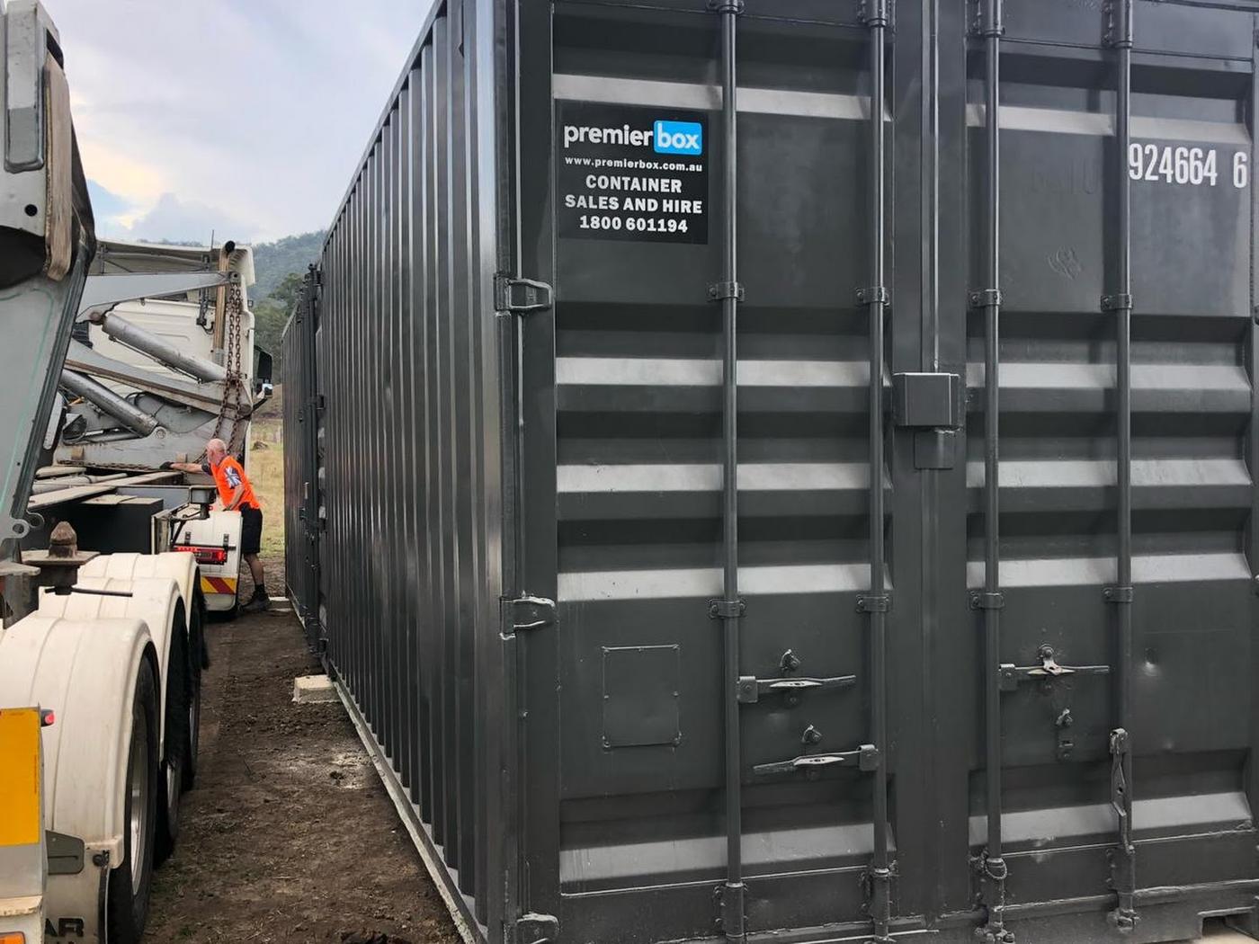 Charcoal Premier Box container beside a side loader truck as the driver sets it down on a rural site