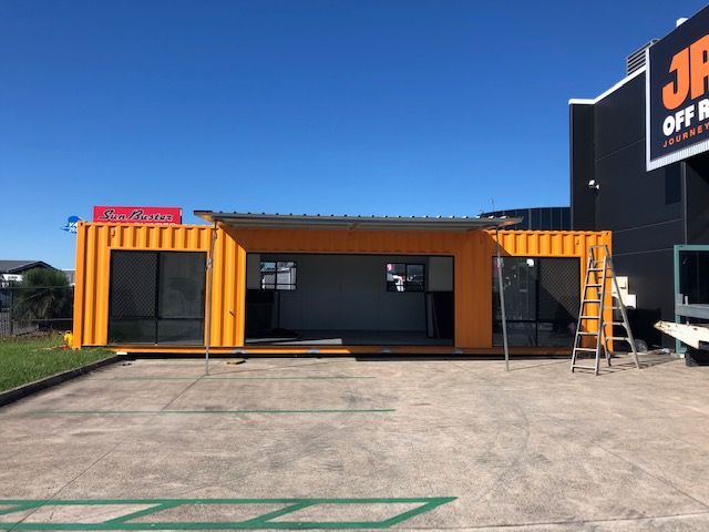 Orange container office with glazed sliding doors and a covered deck beside a showroom